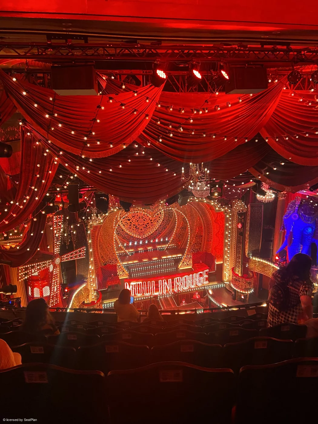 A seat view photo taken from the Grand Circle, showing the stage. On stage there is a red backdrop with hearts, lights and the words 'moulin rouge' written across the centre. At the sides of the image are a windmill and an elephant statue. 