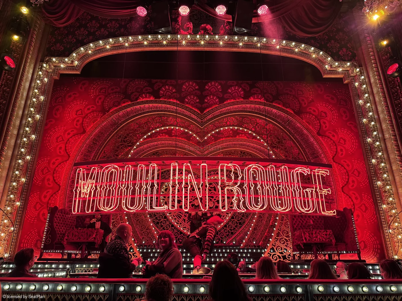 A seat view photo for a best seats moulin rouge blog. It shows the view from the stalls of the stage, a red backdrop with hearts, lights and the words 'moulin rouge' written across the centre.  