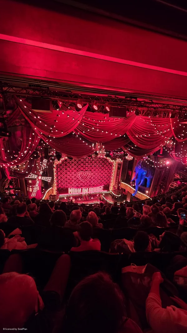 A seat view photo taken from the Grand Circle, showing the stage. On stage there is a red backdrop with hearts, lights and the words 'moulin rouge' written across the centre. At the sides of the image are a windmill and an elephant statue. 