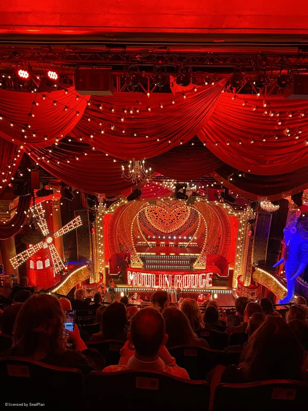 A seat view phototaken from the Grand Circle, showing the stage. On stage there is a red backdrop with hearts, lights and the words 'moulin rouge' written across the centre. At the sides of the image are a windmill and an elephant statue. 
