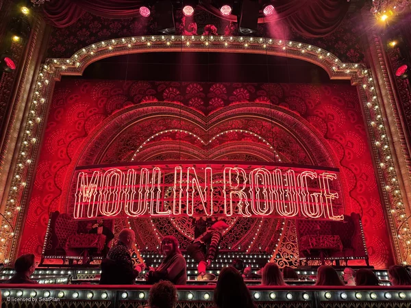 A seat view photo for a best seats moulin rouge blog. It shows the view from the stalls of the stage, a red backdrop with hearts, lights and the words 'moulin rouge' written across the centre.  